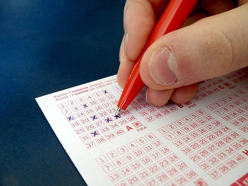 A person holding a red pen is filling out a lottery ticket, marking numbers with blue crosses.