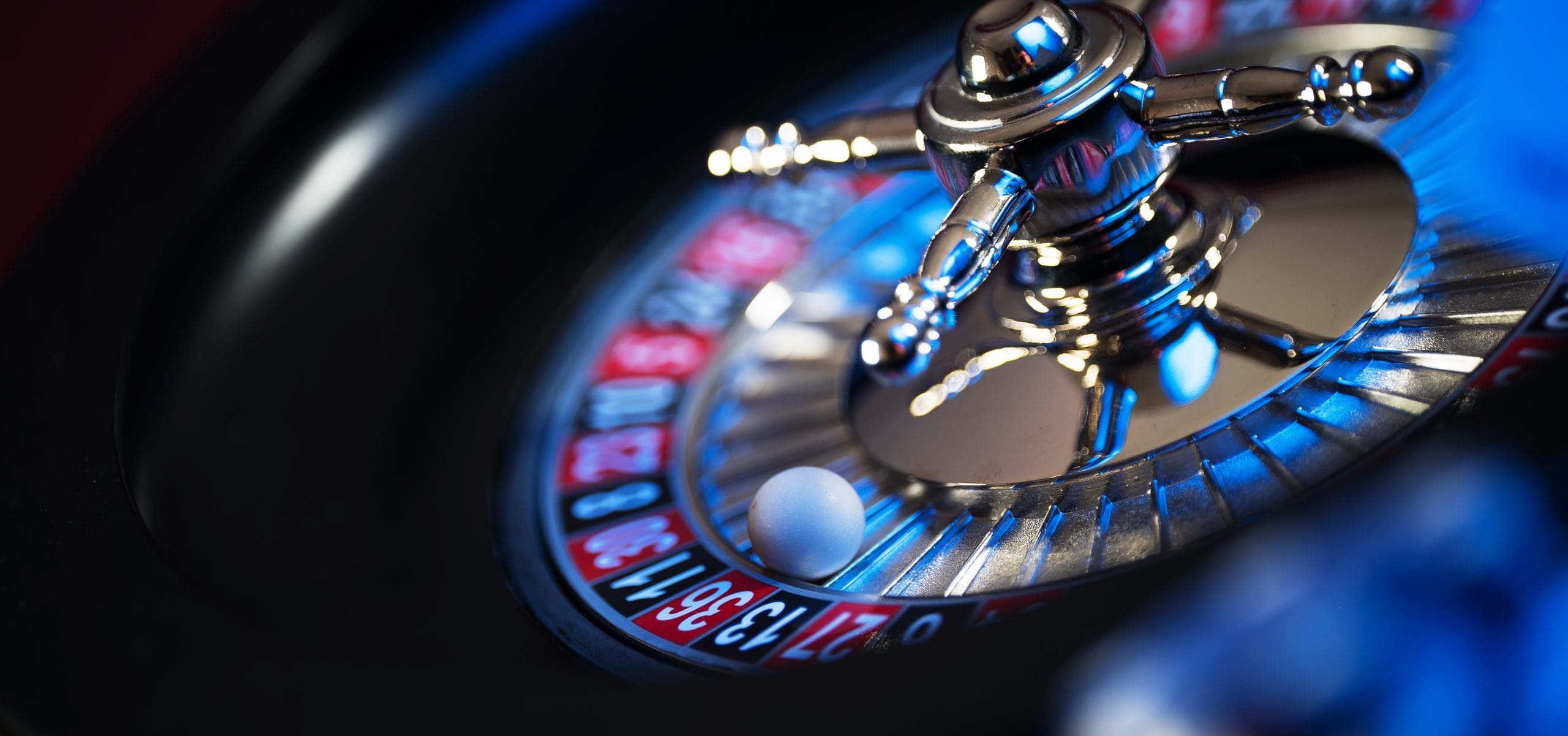 A close-up of a spinning roulette wheel with a white ball resting on the number 21. The wheel features alternating red and black numbered slots, and the image is illuminated with blue lighting.